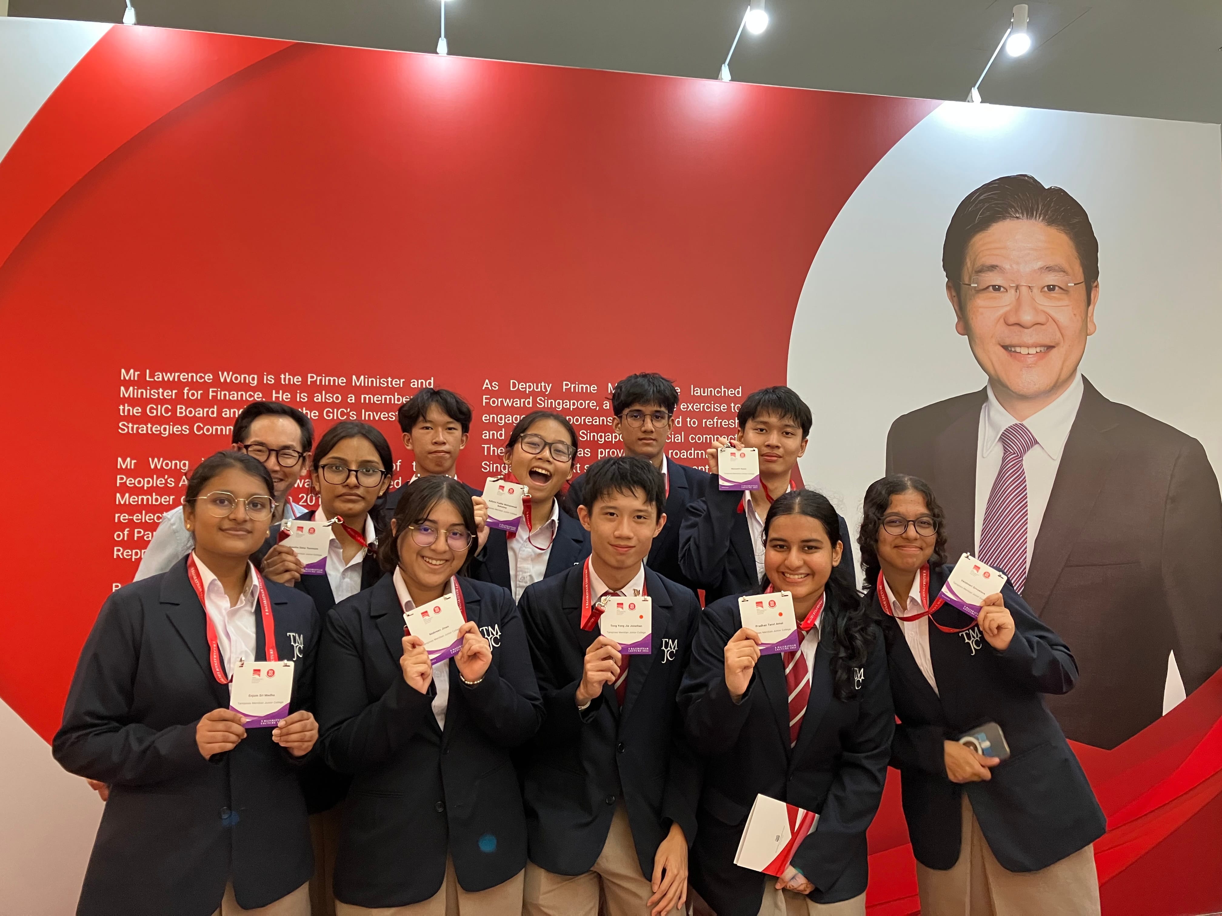 A group of about eleven students in dark blue school blazers pose cheerfully in front of a large red display panel, holding up their event lanyards and name tags. A large portrait and biographical text of Singapore's Prime Minister is visible on the backdrop behind them.
